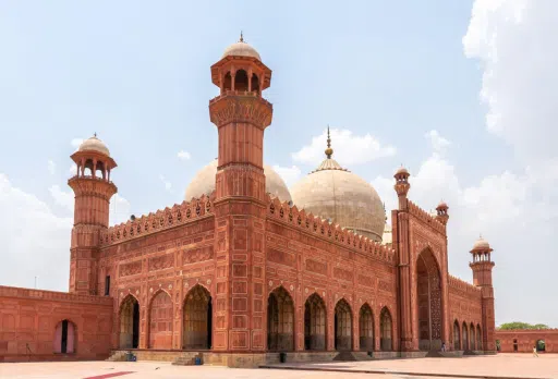 A large, historic mosque with red stone architecture and multiple domes, supported by Giving Hands.