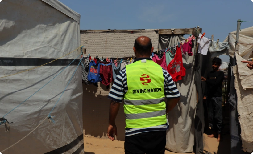 Giving Hands كندا aid worker providing humanitarian support in a Gaza refugee camp with tents and laundry hanging.